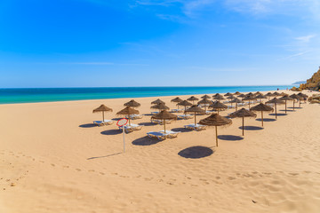 Sunbeds with umbrellas on sandy beach in Salema seaside town, Alrgarve region, Portugal