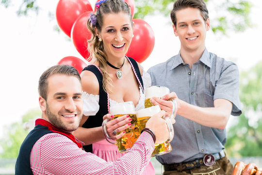 Drei Freunde in M&uuml;nchner Biergarten in Tracht sto&szlig;en mit Hellem an