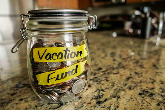 Vacation Fund Money Jar. A Clear Glass Jar Filed With Coins And Bills, Saving Money. The Words 