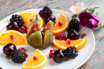 Healthy breakfast of fresh fruit including fig, sliced orange, blackberries, cherries and pomegranate seeds. Served on a rustic wooden table with fresh wild flowers.