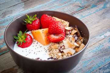 A bowl of healthy breakfast muesli with yogurt and topped with fresh strawberries, sliced melon, goji berries, carob and coconut shavings.