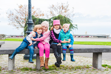 4 cute kids sitting in the bench in a city, wearing warm waistcoats and boots