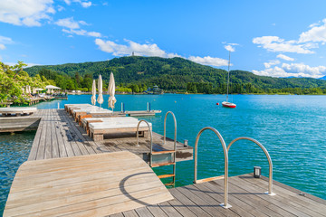 Obraz premium Sunchairs on wooden deck and view of beautiful alpine lake Worthersee in summer, Austria