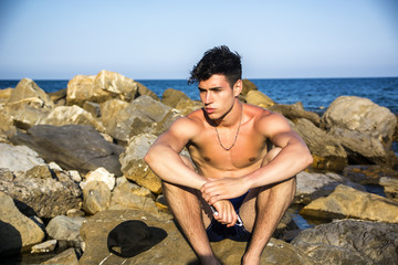 Young shirtless athletic man sitting on rock by sea