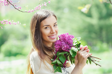 Fototapeta premium Happy lifestyle concept with attractive smiling young woman holding a bunch of spring blooming lilac close to her face