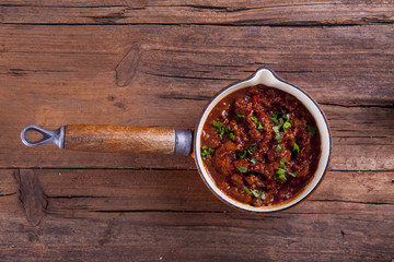 Mince meat ragu with chopped herbs on shot in a pot on a wooden background from above