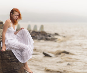 Redhead woman sitting on the coast on beach and looks into the distance at sea. Selective soft focus.