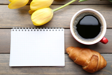 Cup of coffee with fresh croissant, tulips and blank sheet of paper on wooden background