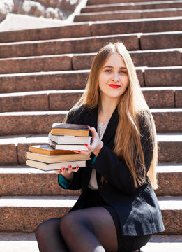 Beautiful girl student is sitting on stairs with stack of books