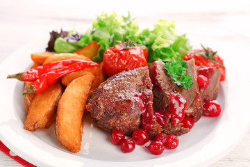 Tasty roasted meat with cranberry sauce and roasted vegetables on plate, on color wooden background