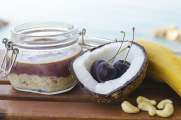 Cherry coconut banana overnight oats with cracked open coco, banana, cashews mixed with chia seeds and cherry cream in a mason jar, on a tropical sea background. Healthy fitness breakfast oatmeal.