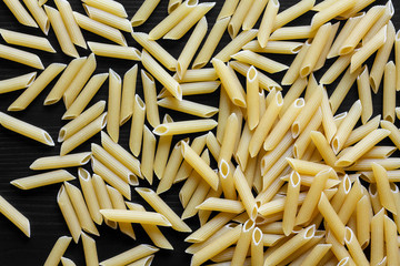 Dry penne pasta on black textured wood from above.