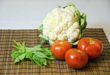 A raw cauliflower vegetable, three tomatoes and a bunch of fresh organic basil on a rustic bamboo tablecloth