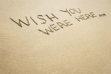 Words wish you were here written in the sand on the beach.