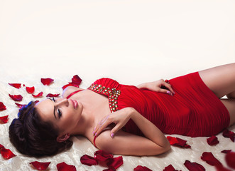 Obraz na płótnie Canvas Sexy beautiful brunette woman in red dress laying on a bed under romantic falling rose petals 