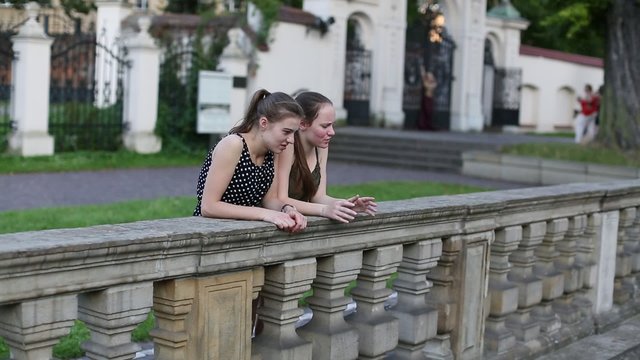 Two young cute girls standing near the stone fence, spend time talking.