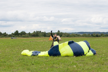 Cheerful parachutist in black suit with striped yellow-blue parachute on green field