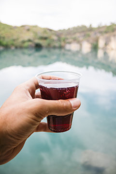 A Hand Of A Man Holding A Glass Of Soda In Front Of A Lake