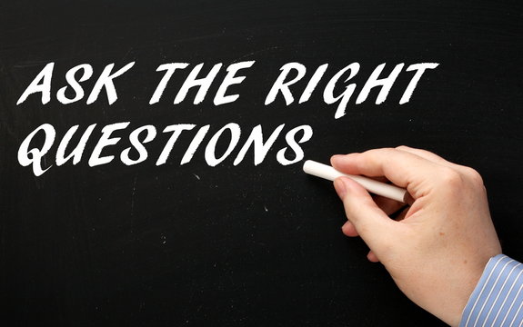Male hand wearing a business shirt writing the phrase Ask The Right Questions in white text on a blackboard