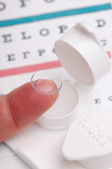 Contact Lens and Case &ndash; A contact lens on a finger. Contact lens case and eye chart in background.