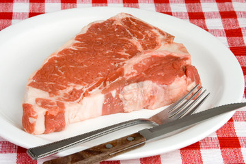 Raw Steak on White Plate – A raw T-bone steak on a white place. Red checked background.