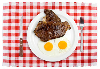T-Bone Steak and Fried Eggs Breakfast – Two sunny side up eggs and a T-Bone steak on a white plate. Table setting, with fork and steak knife. Red checked background.