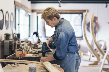 Elderly woman artist drilling wood in her workshop
