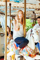 Fototapeta na wymiar On a summer day a group of friends having fun in the shade on a carousel with horses and blowing soap bubbles. In the foreground a young woman in the air while blowing many colorful soap bubbles