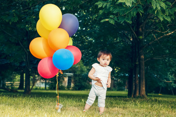 Fototapeta na wymiar Portrait of cute adorable little asian girl child one years old, in white pants shirt, standing with balloons on field meadow on sunset, concept of happy birthday holiday, toned with Instagram filters