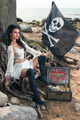 Beautiful pirate woman sitting near treasure chest on the beach with a bottle of rum and classic...