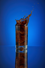 Splash of cola soft drink on a blue background. Refreshing  liquid drink coca pouring into a glass with ice. Pour high speed beverage for promoting restaurant and bar.  isolated liquor.
