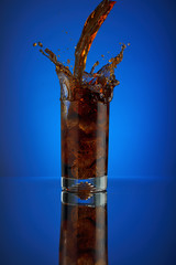 Splash of cola soft drink on a blue background. Refreshing  liquid drink coca pouring into a glass with ice. Pour high speed beverage for promoting restaurant and bar.  isolated design liquor.
