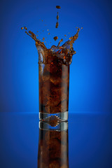 Splash of cola soft drink on a blue background. Refreshing  liquid drink coca pouring into a glass with ice. Pour high speed beverage for promoting restaurant and bar. 