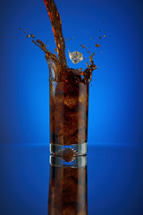Refreshing splash of cola soft drink on a blue background. Liquid drink coca pouring into a glass with ice. Pour high speed beverage for promoting restaurant and bar. Closeup isolated design liquor.
