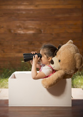 Little girl with binoculars inside white wooden box with toy bear exploring surroundings