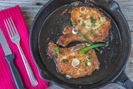 horizontal image of two pork chops fried to a golden brown in a black cast iron pan with  green onion sprinkled all over the top with a fork and knife lying on a red napkin on a rustic wood background