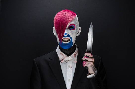 Clown and Halloween theme: Scary clown with pink hair in a black suit with a knife in his hand on a dark background in the studio
