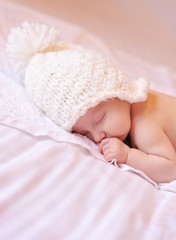 Little baby sleeping on the bed with white cover and knitted white hat on. Childhood. 