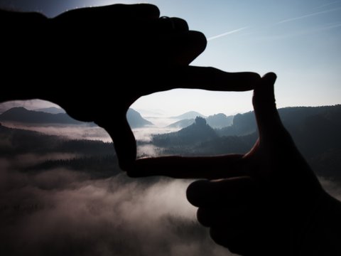 Fingers frame. Close up of hands making frame gesture. Blue misty valley bellow rocky peak. Sunny spring in rocky mountains.