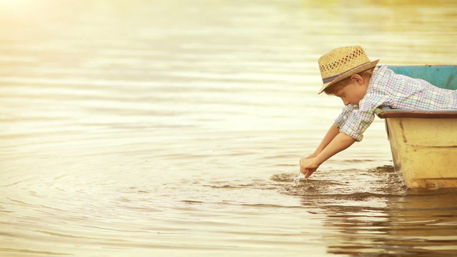 Warmly toned golden footage: wee boy in a trendy hat dangles his hands in the water being in the boat himself.