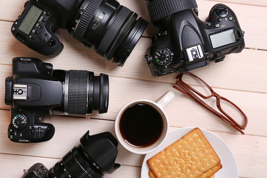 Modern cameras with glasses, cup of coffee and cookies on wooden table, top view