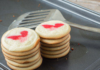 horizontal close up image of two stacks of freshly baked warm cookies with heart shape pattern on top lying in a cookie sheet with spatula beside it.