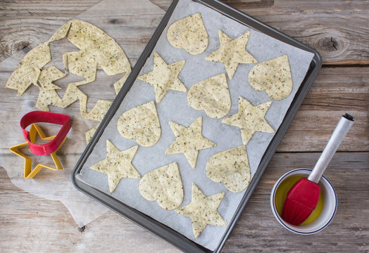 horizontal image of the process of the makings of tortilla chips made into heart and star shapes lying in cookie sheet with some left over tortilla dough and a cookie cutter on rustic wood surface.