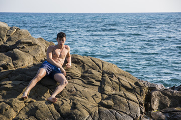 Fototapeta na wymiar Young shirtless athletic man leaning on rock by water on ocean