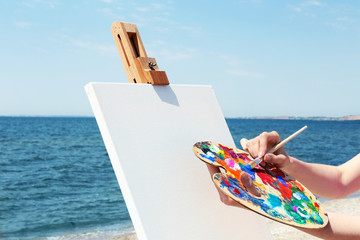 Female hand holding palette and easel with canvas on beach