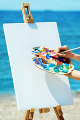 Female hand holding palette with paints and easel with canvas on beach