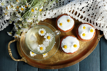 Cup of chamomile tea with chamomile flowers and tasty muffins on tray, on color wooden background