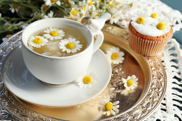 Cup of chamomile tea with chamomile flowers and tasty muffins on tray, on color wooden background
