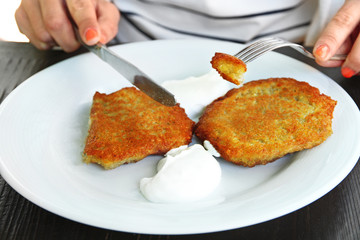Female hands cutting potato pancakes with cream in plate on wooden table, closeup