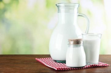 Pitcher, jar and glass of milk on wooden table, on nature background